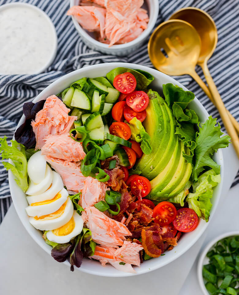 Cobb salad in a bowl made up of salmon pieces, tomatoes, avocado slices, greens and slices of hard boiled egg