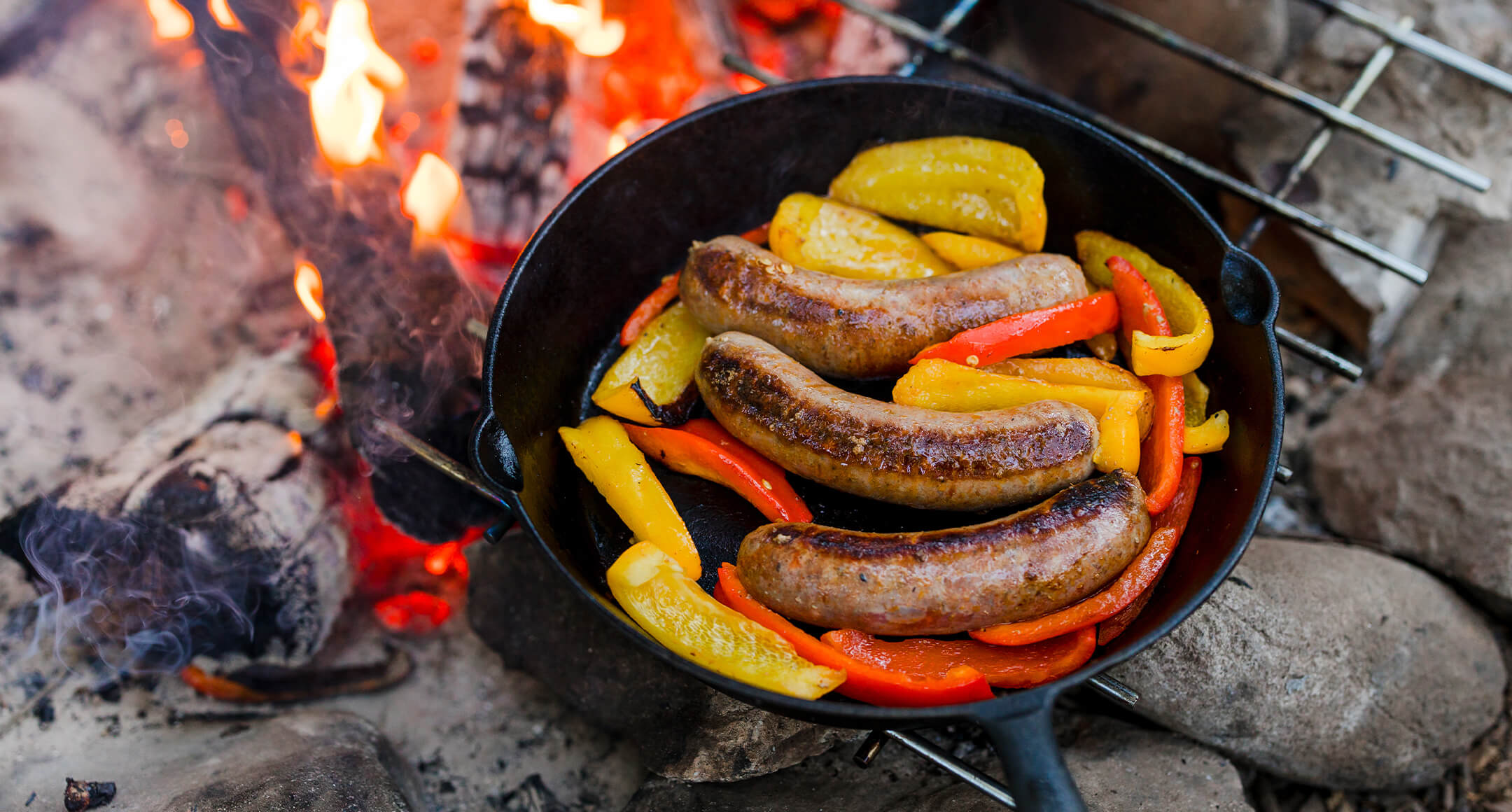 Sausages and bell peppers roasting in a cast-iron pan on a grill grate over a campfire.  