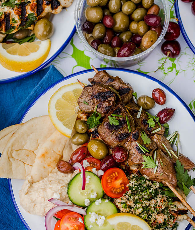 an outdoor dining table scene featuring a plate with kebabs, pita, hummus, olives, taboulleh salad, cucumbers, tomatoes, onions, sliced lemons and herbs.