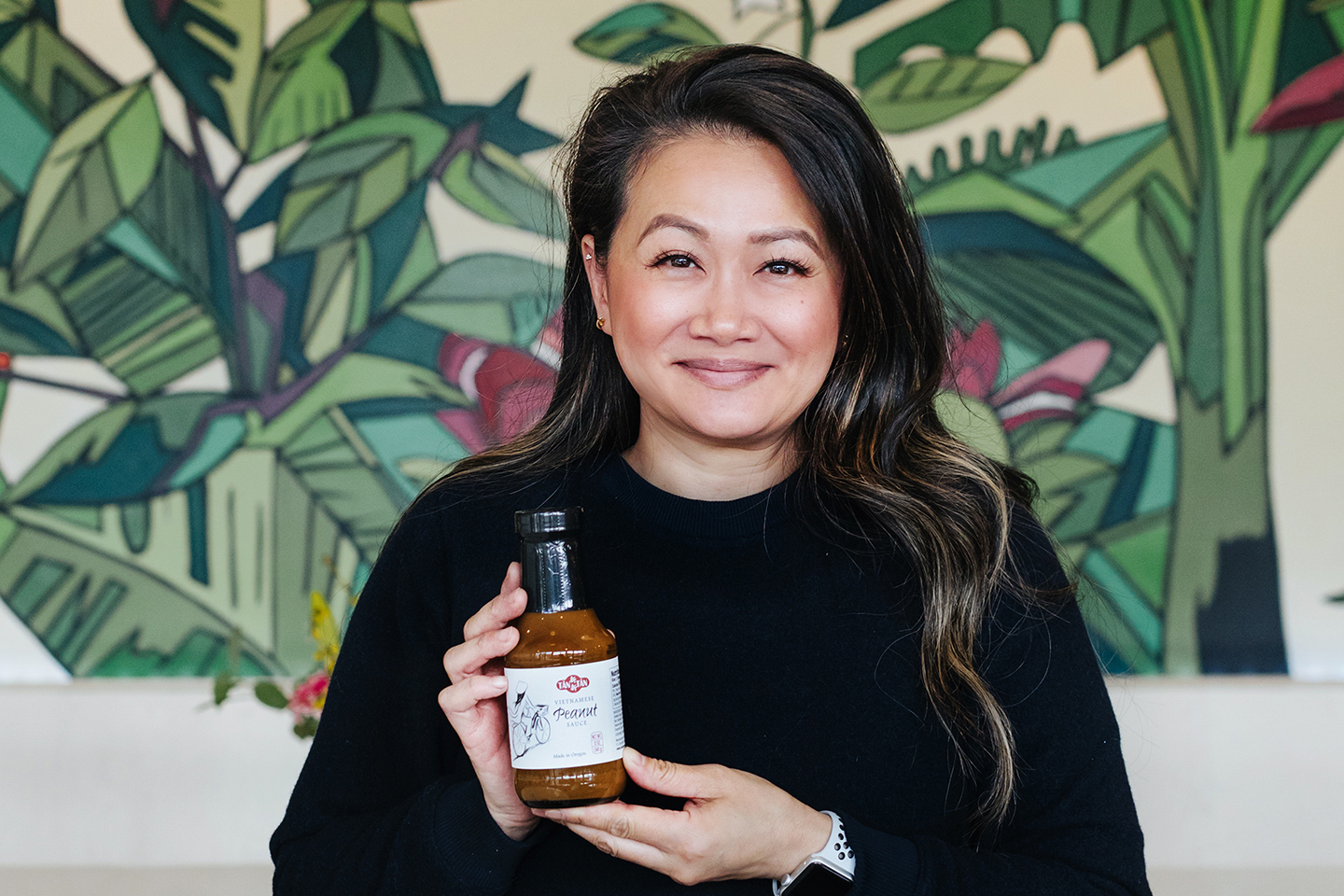 Smiling women holding her product Tan Tan Vietnamese Peanut Sauce in front of a tropical mural.