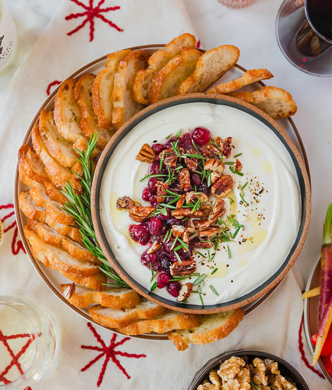 a beautifully curated holiday appetizer dip topped with cranberries and nuts and surrounded by sliced baguette