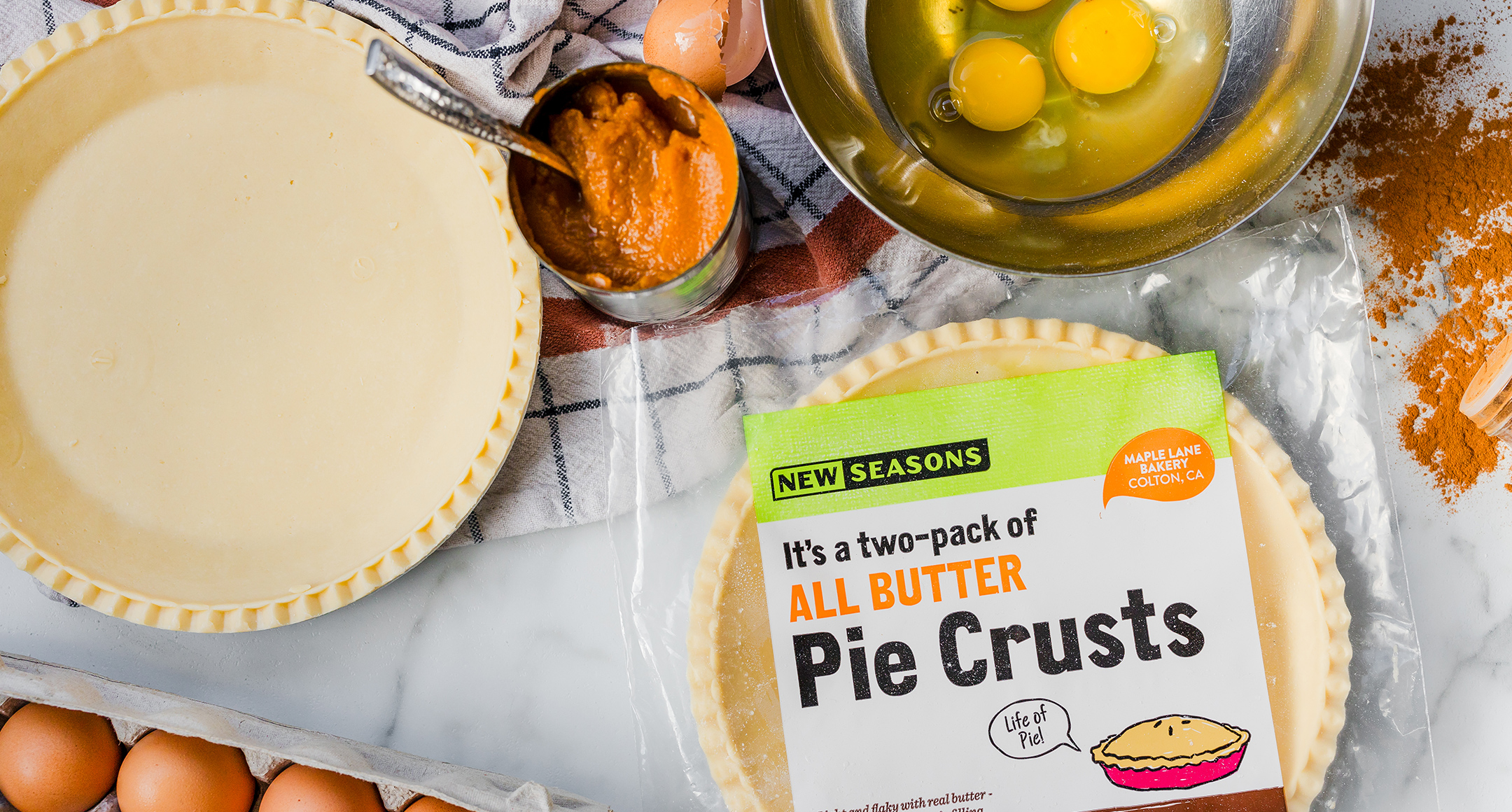 A countertop spread featuring the ingredients of a pumpkin pie, including pie crust, eggs, spices, and a can of pumpkin purée. 