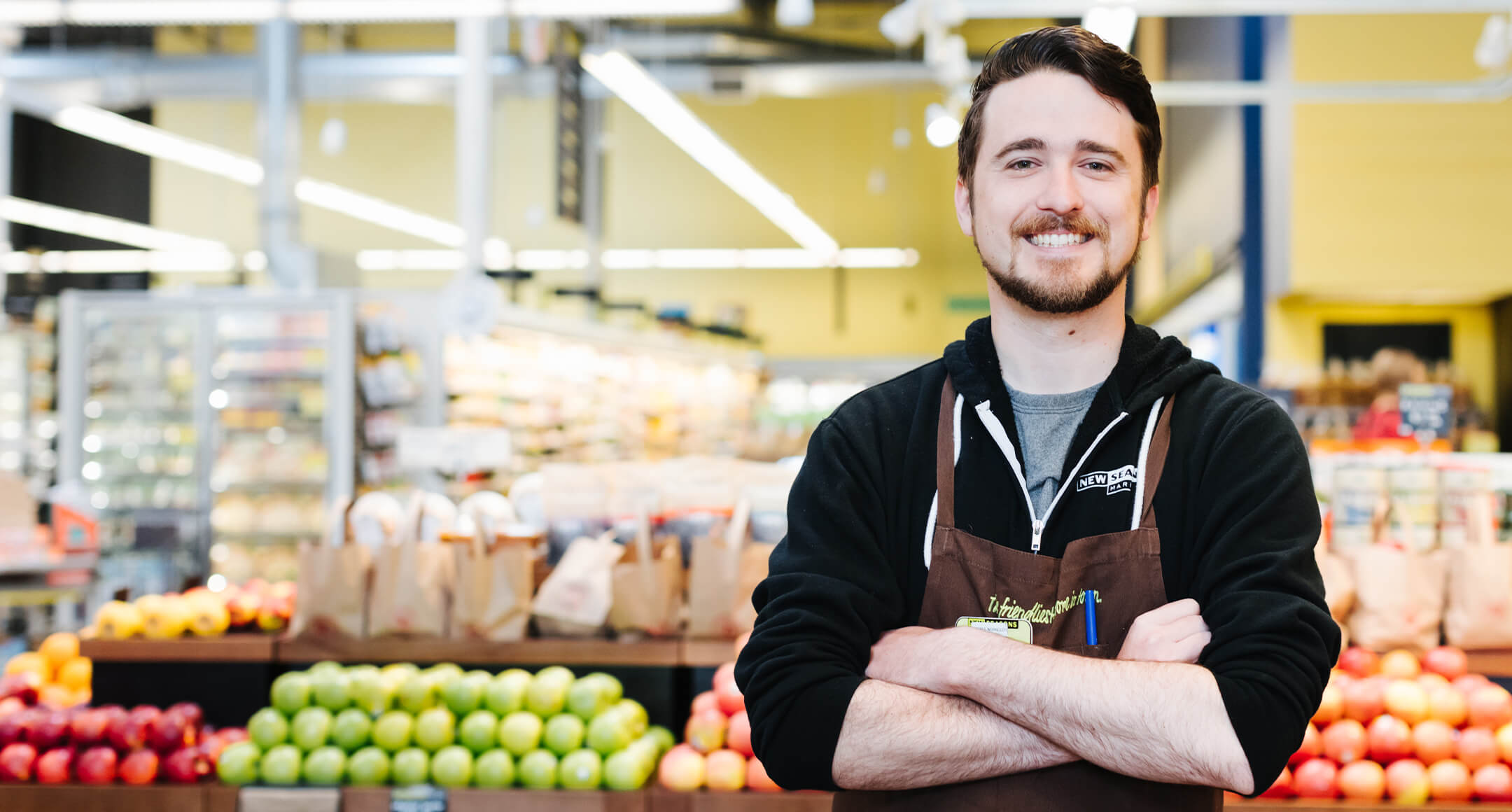 Smiling staff member at New Seasons Market checkout standing in front of apple displays in produce.