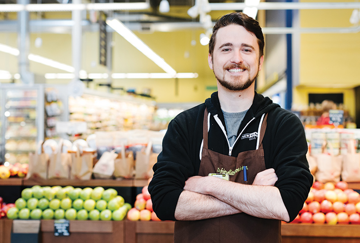 Smiling staff member at New Seasons Market checkout standing in front of apple displays in produce.