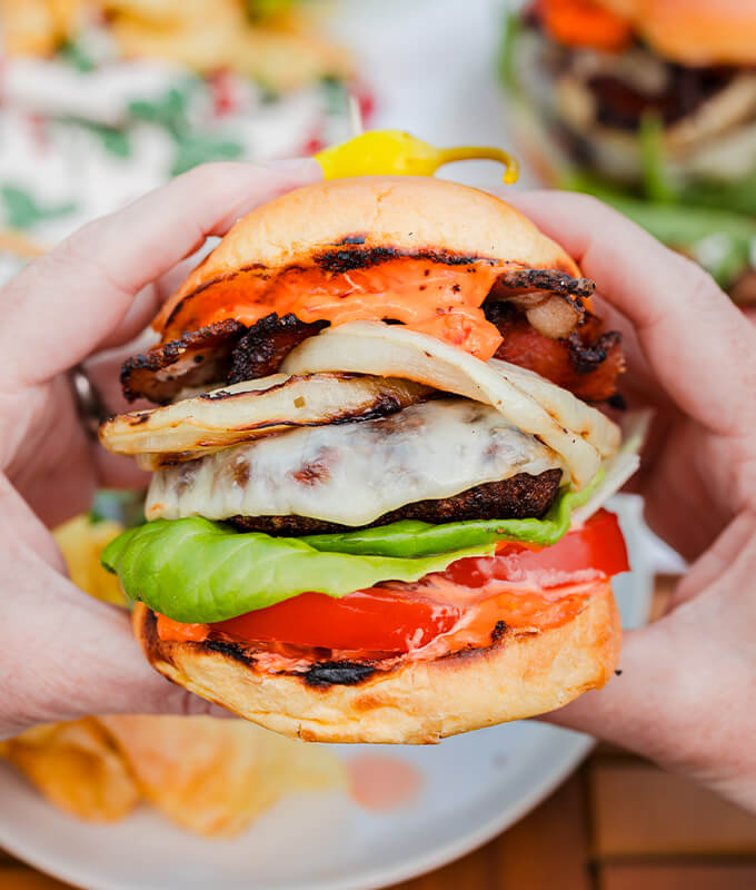 An image of two hands holding a burger with lettuce, cheese, tomatoes, onions, and a pepper on top. 