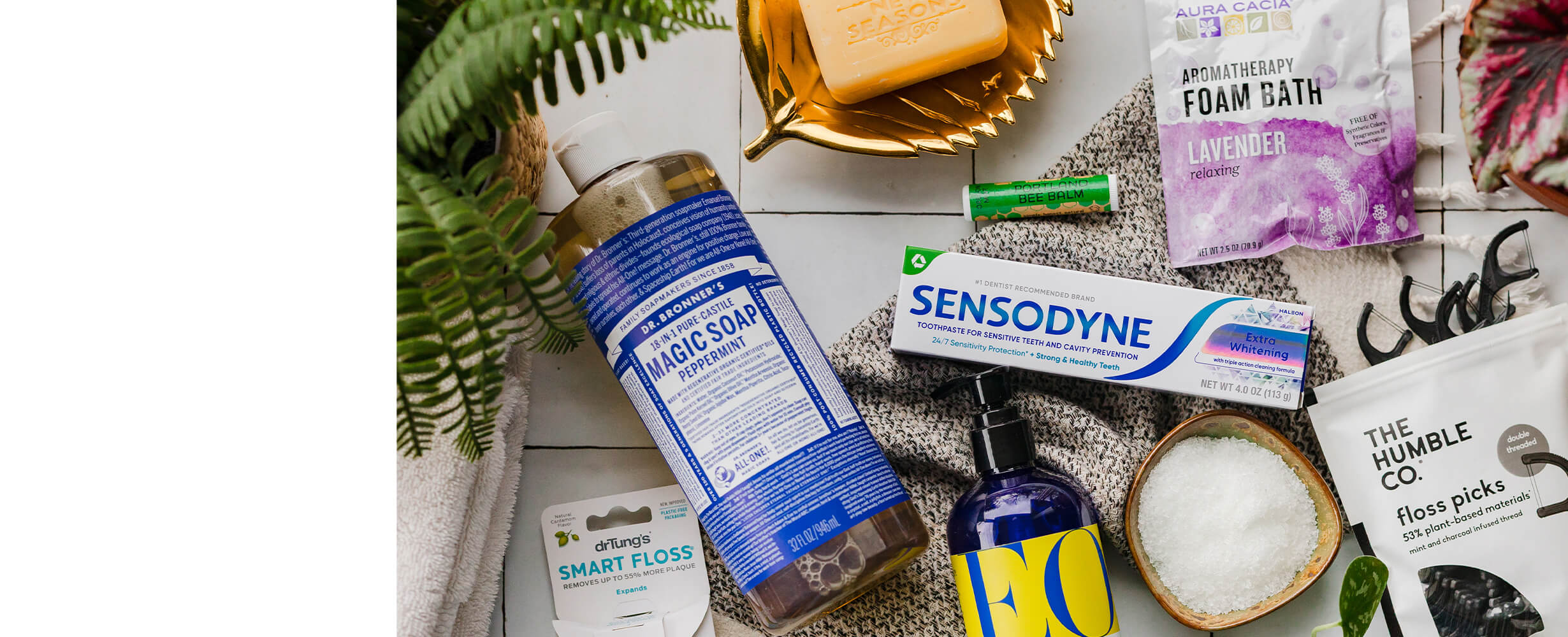 A top down image of an assortment of body care products including Dr. Bronner's Magic Soap, Sensodyne toothpaste, Aura Cacia Aromatherapy Foam Bath, Portland Bee Balm, and The Humble Co Floss Picks.