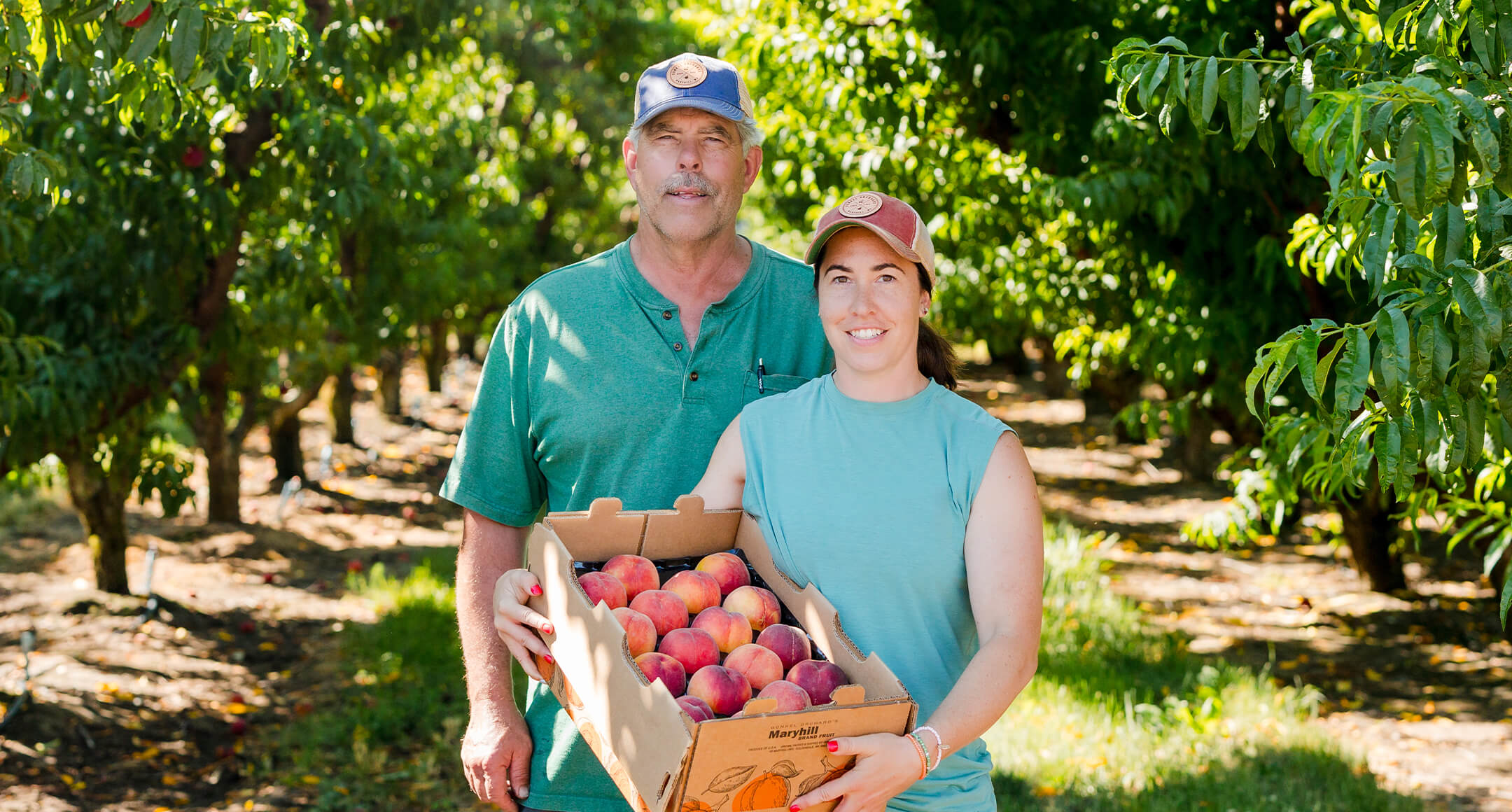Two members of the Gunkels smiling and holding a box of peaches in their orchard. 