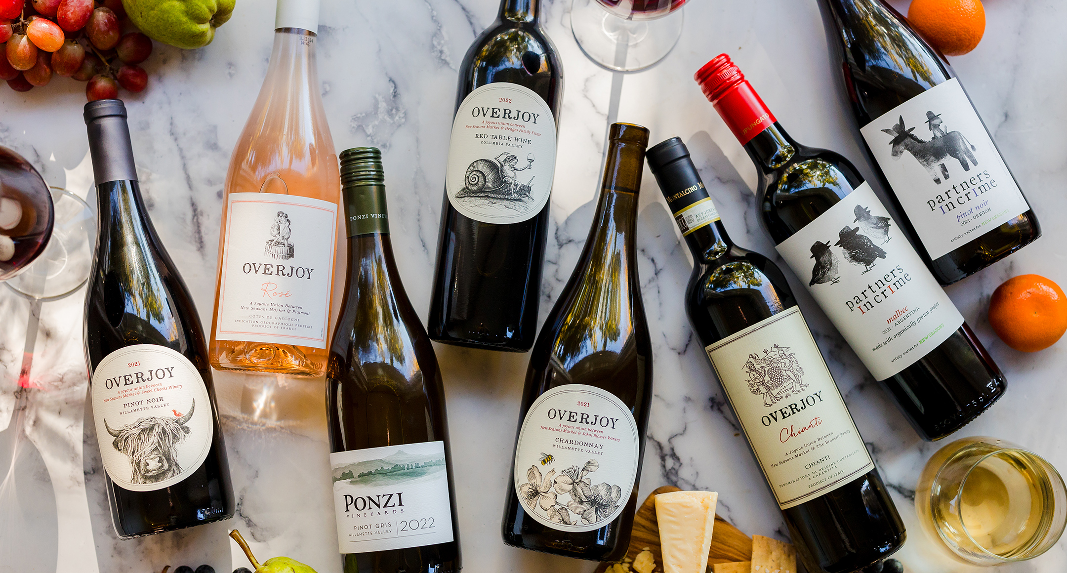 A wide selection of white and red wine bottles displayed side by side on a counter with grapes, oranges, and filled wine glasses. 