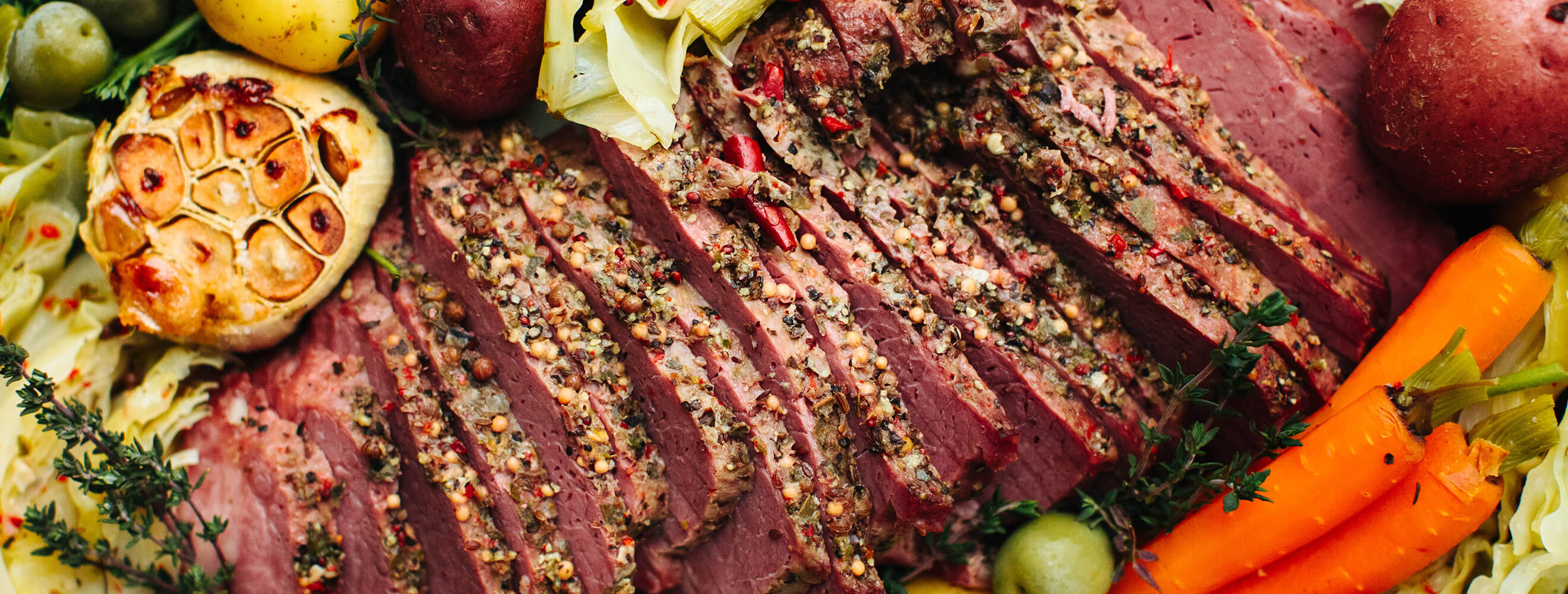 A top down image of a seasoned cooked corned beef in a bed of vegetables including garlic, carrots, and potatoes.