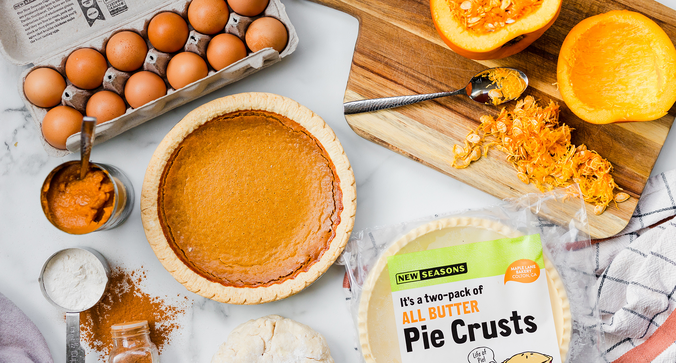 A freshly baked pumpkin pie on a countertop along with the ingredients it was made from, including a carton of eggs, a fresh pumpkin cut to expose the inside, a can of pumpkin purée, a store-bought pie crust, and spices.