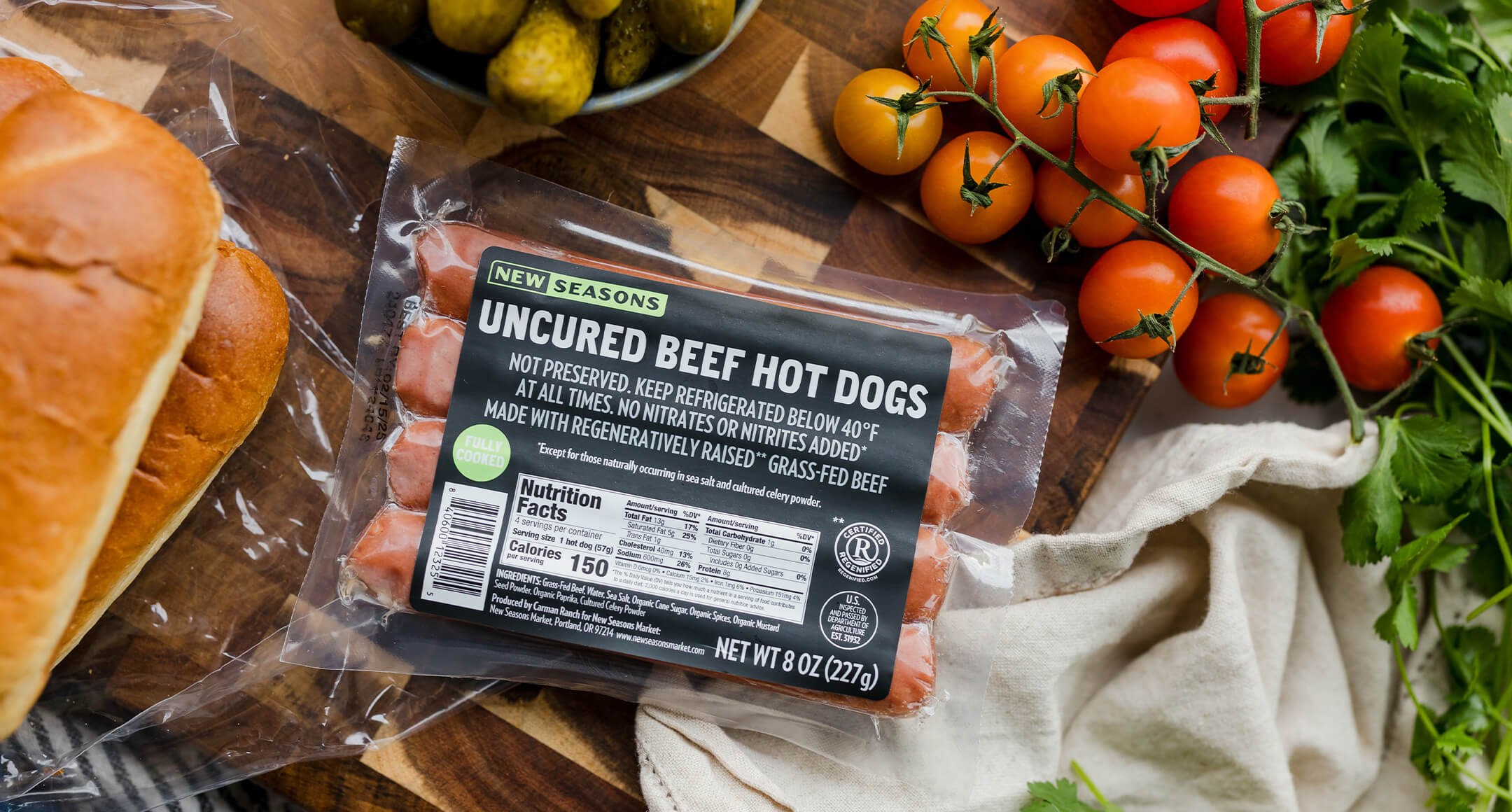 Package of New Seasons Market uncured beef hot dogs arranged on a cutting board with buns and tomatoes on the vine.